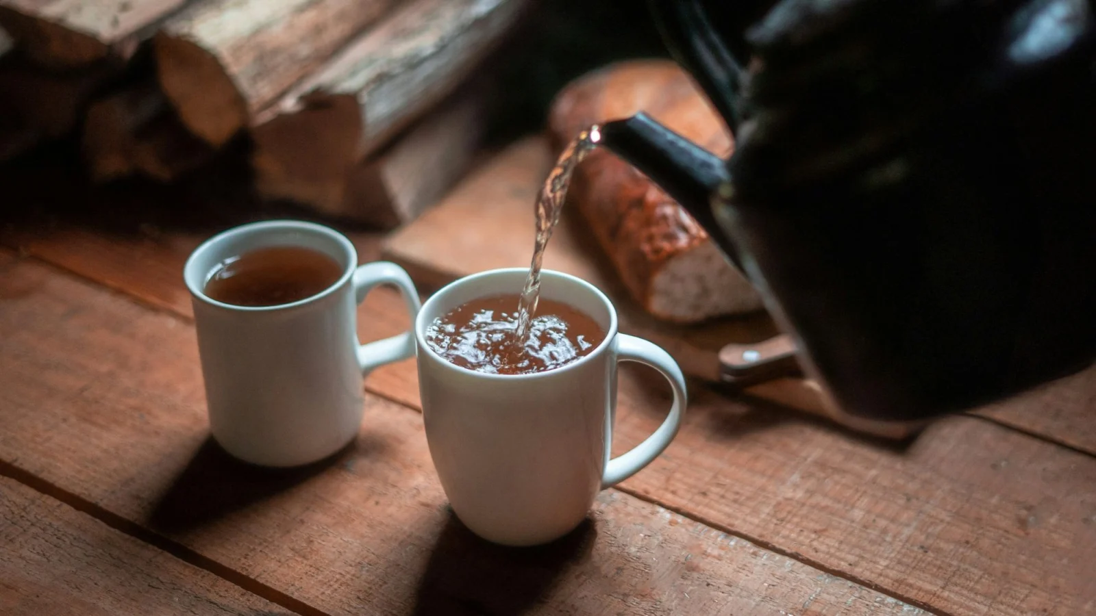 A black kettle pouring hot water into two mugs of black tea on a wooden table
