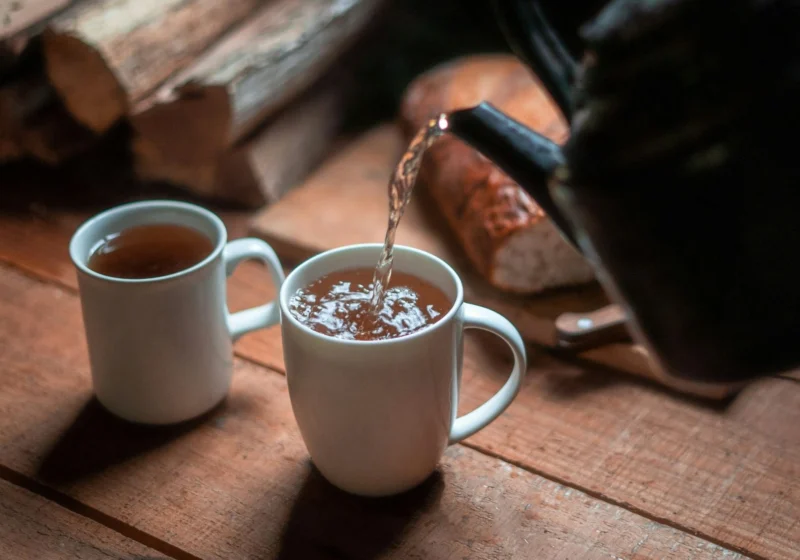 A black kettle pouring hot water into two mugs of black tea on a wooden table
