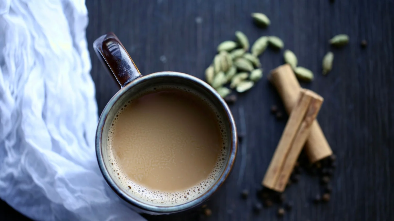 A mug of milk tea with green cardamom pods and cinnamon on dark wood