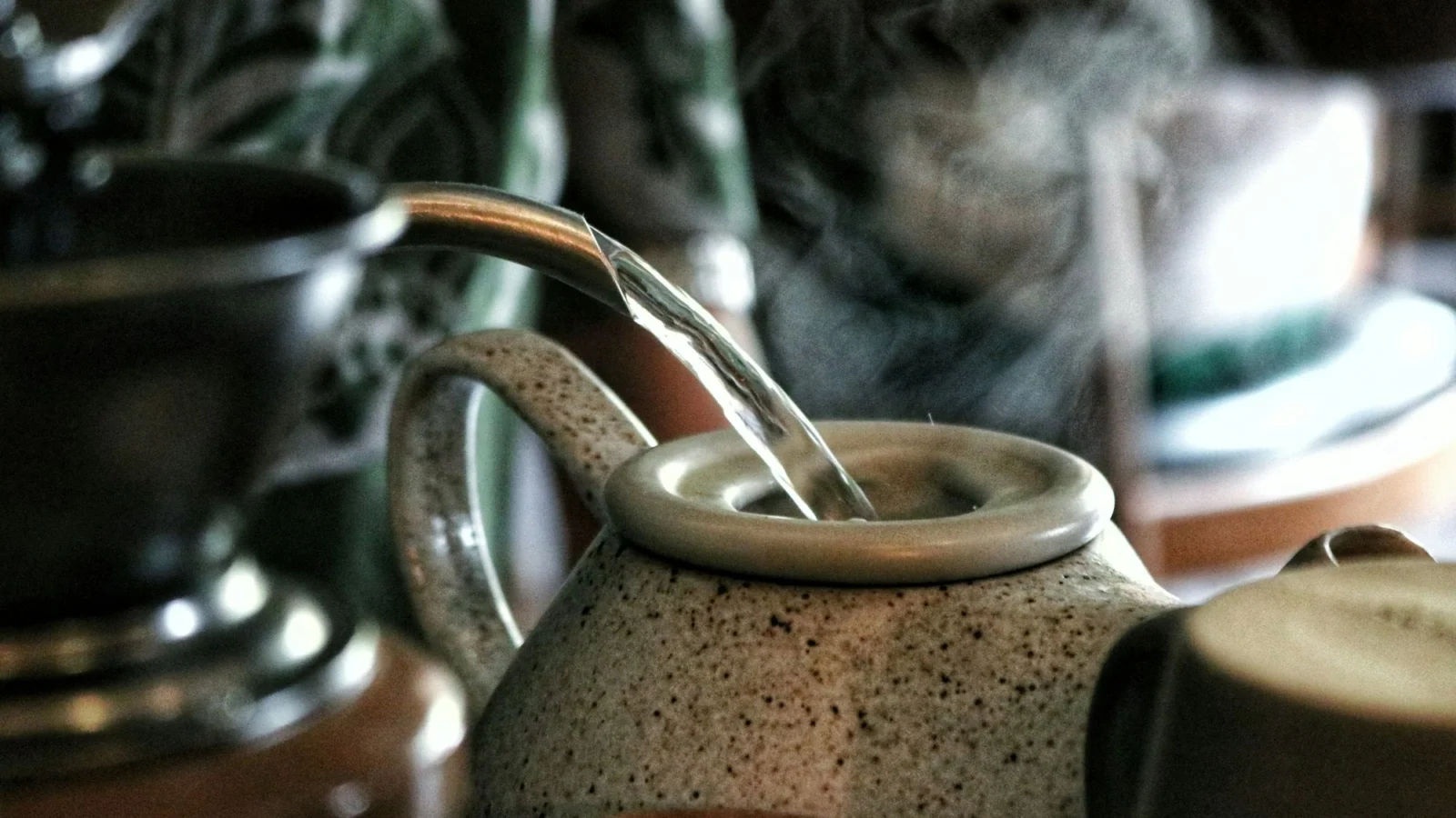 A kettle pouring steaming water into a stoneware teapot
