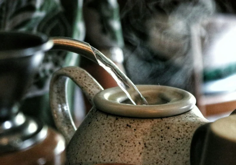 A kettle pouring steaming water into a stoneware teapot