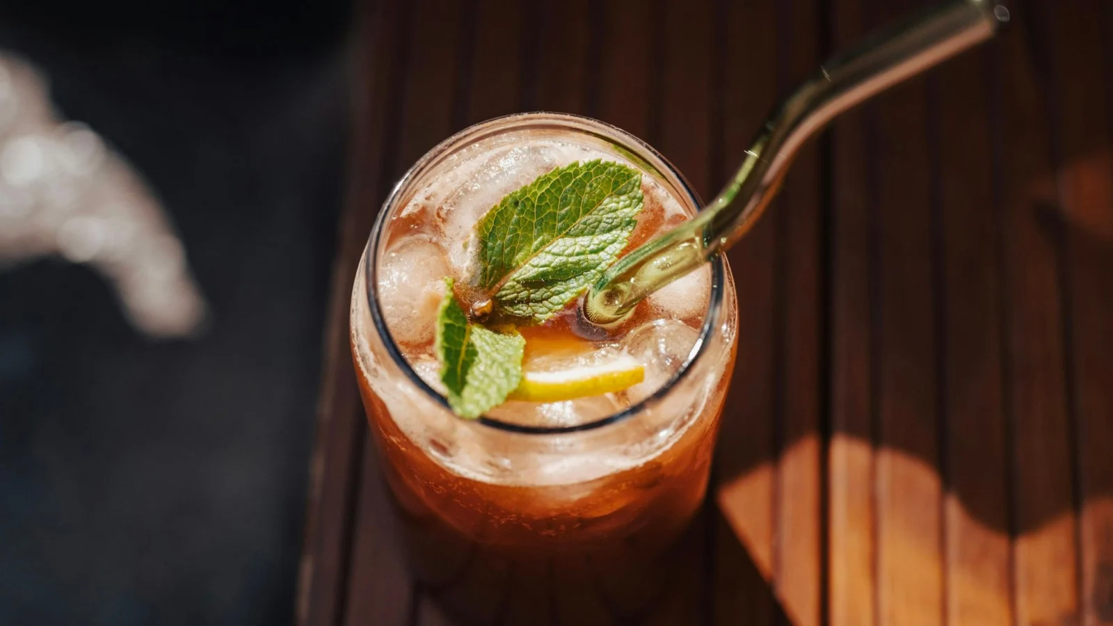 A glass of sparkling iced tea with lemon, mint and a glass straw on dark wood
