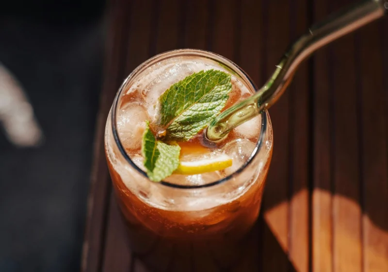 A glass of sparkling iced tea with lemon, mint and a glass straw on dark wood