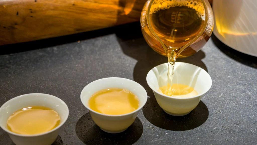 Golden tea being poured from a glass teapot into small white cups